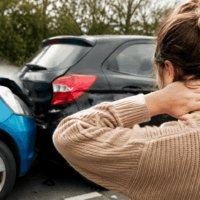 Person in a beige knit sweater holds their neck in front of a blue car and a black car that have collided on the road, suggesting a crash scene. Person in a beige knit sweater holds their neck in front of a blue car and a black car that have collided on the road, suggesting a crash scene.